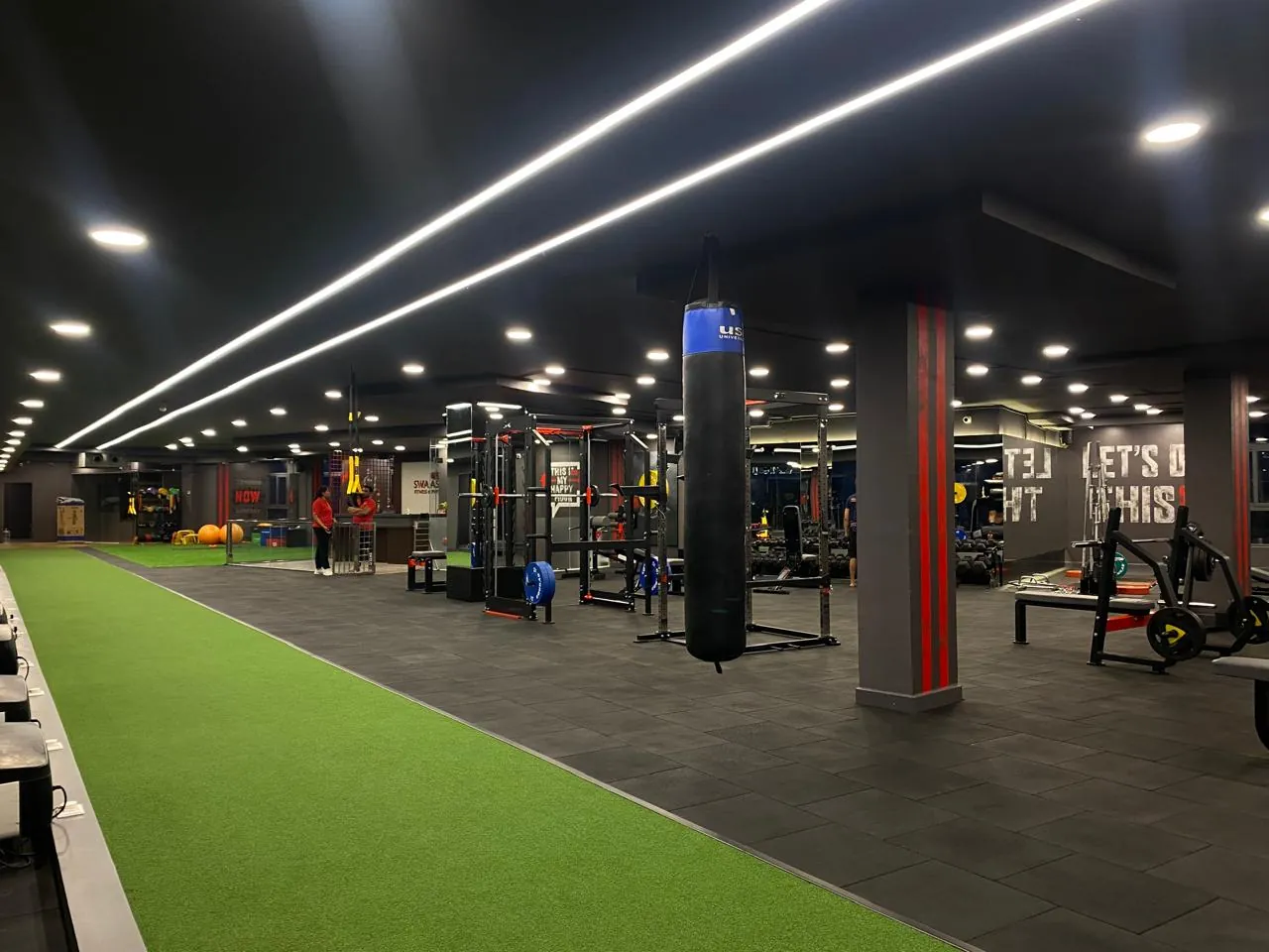 The Swaasthya gym floor with power racks, a punching bag and a turf track under strip lighting